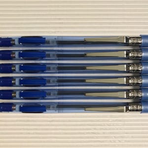 以下が含まれることがあります： 青と透明のボールペン6本のコレクション。ペンは透明なバレル、青インク、銀色の金属アクセントが付いています。明るい色の表面に平行に配置されています。