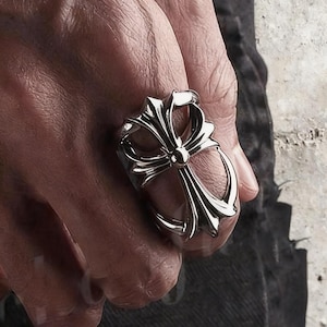 May include: A silver-toned metal ring featuring a prominent cross design with curved, flared arms and a central circular detail. The ring has a polished, reflective finish and is worn on the finger, showcasing its intricate, symmetrical metalwork.