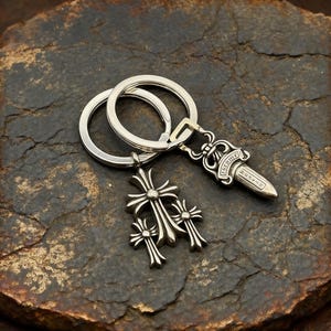 May include: Silver-colored keychain with two rings, featuring a dagger pendant with the word "Chrome" and a cross pendant with three crosses. The keychain is displayed on a dark, textured surface.