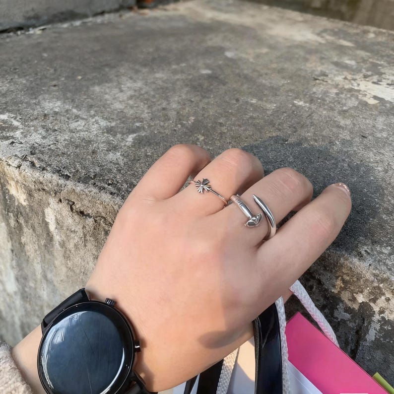May include: A silver ring featuring a small cross emblem and a second silver ring with a curved, open-ended design. A black wristwatch with a round face and a black strap is worn on the wrist, accompanied by a partial view of a shopping bag.