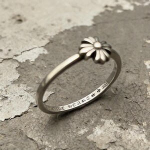 May include: A silver-toned ring featuring a small, multi-petaled flower motif at the top. The interior of the band is engraved with the text CHROME HEARTS, a small cross symbol, and the number 2011 in a decorative, gothic-style typeface.