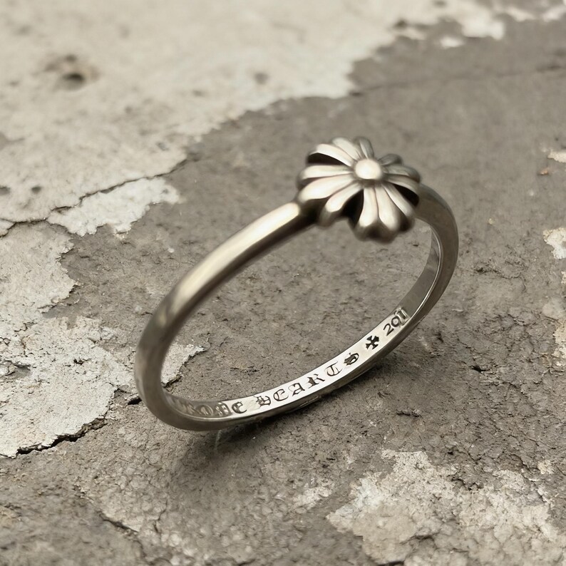 May include: A silver-toned ring featuring a small, multi-petaled flower motif at the top. The interior of the band is engraved with the text CHROME HEARTS, a small cross symbol, and the number 2011 in a decorative, gothic-style typeface.