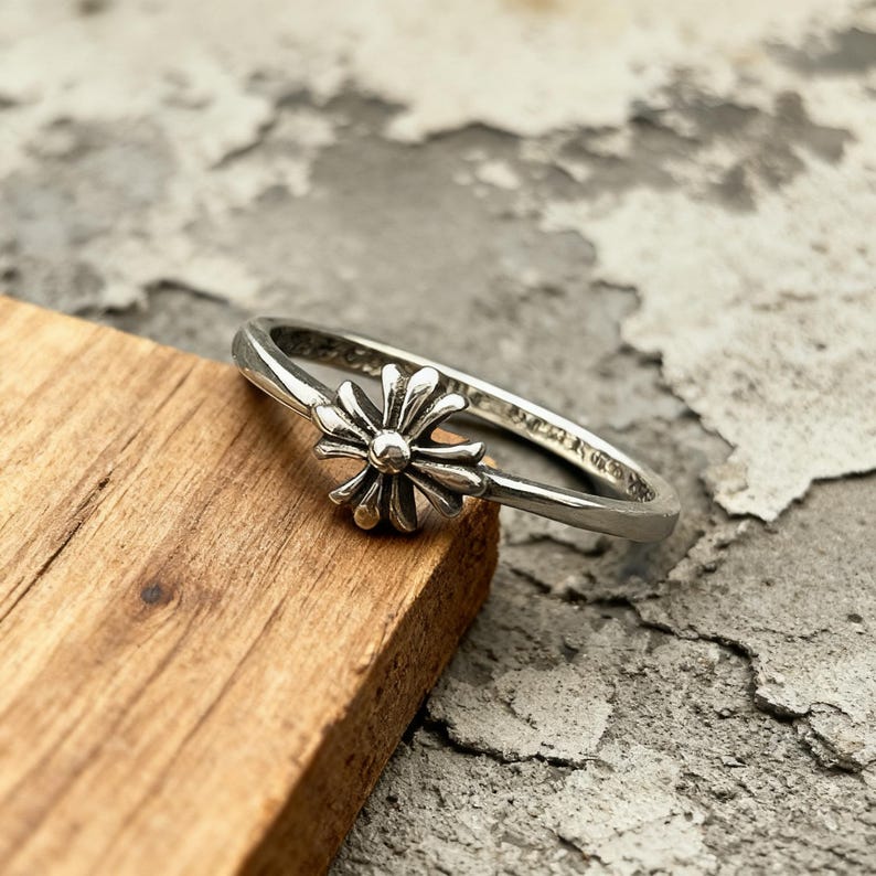 May include: A silver ring featuring a central floral-shaped motif with multiple petals radiating from a small circular centre. The band is thin and polished, showcasing a metallic finish with darkened recessed areas that highlight the intricate design.