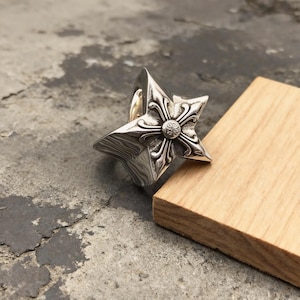 May include: A silver ring shaped like a five-pointed star with a polished finish. The center of the star features an embossed cross design with intricate, curved detailing and a small circular element at the very middle of the cross.