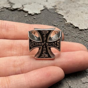 May include: A silver-toned metal ring featuring a large iron cross design. The surface of the cross is black with silver lettering that reads "Enfants Riches" in a gothic-style font. The ring has a polished finish and a wide, prominent face.