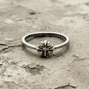 May include: A silver ring featuring a central cross emblem with rounded, petal-like arms. The band is thin and polished, with engraved text visible along the inner circumference of the metal ring, showcasing a detailed and metallic finish throughout.