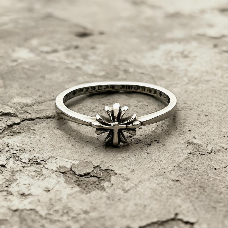 May include: A silver ring featuring a central cross emblem with rounded, petal-like arms. The band is thin and polished, with engraved text visible along the inner circumference of the metal ring, showcasing a detailed and metallic finish throughout.