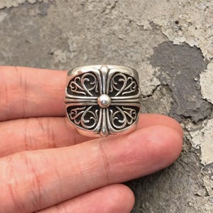 May include: A wide silver ring featuring a prominent central cross motif with intricate, symmetrical scrollwork patterns engraved into the metal surface. The design includes a small, smooth, raised circular element at the very center of the cross.