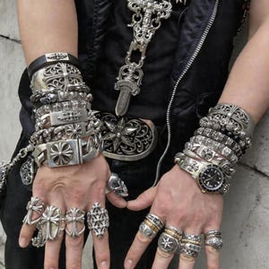 Pode incluir: Uma coleção de joias prateadas, incluindo várias pulseiras sobrepostas, um relógio de pulso com mostrador preto e numerosos anéis com motivos de cruz e florais. Uma grande fivela de cinto ornamentada e um longo pingente são visíveis.