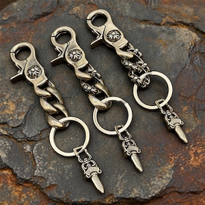 May include: Three antique silver keychains with ornate details. Each keychain features a clasp, a chain, a ring, and a decorative dagger pendant. The keychains are displayed on a dark, textured surface.