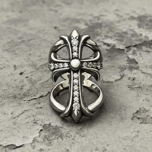 Könnte beinhalten: Ein silberfarbener Ring mit einem markanten Kreuzdesign. Das Kreuz ist mit einer vertikalen Reihe kleiner, klarer, funkelnder Steine und einem zentralen runden Stein verziert. Der Metallring hat eine polierte Oberfläche mit geschwungenen Details.
