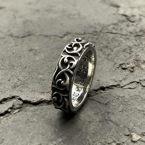 May include: A silver ring featuring an intricate, raised scrollwork pattern across its outer band. The interior of the band is engraved with the text "RUS 2000 925". The metal has a polished finish with darkened recessed areas to highlight the design.