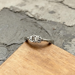 Könnte beinhalten: Ein silberfarbener Metallring mit einem markanten, erhabenen gotischen Buchstaben-Design auf der Vorderseite. Der Ring ist schmal und poliert und weist eine metallische Oberfläche mit komplexen Details auf dem zentralen Emblem auf.