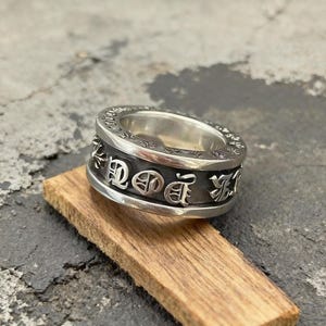 May include: A silver ring featuring a wide band with a dark, recessed centre section. The band is decorated with raised, polished silver gothic-style lettering and small cross motifs. The edges of the ring are smooth, polished, and rounded.