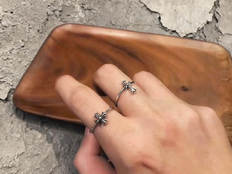 May include: Two silver rings featuring a detailed cross emblem on the band. The rings have a polished metallic finish with darkened recessed areas to highlight the intricate cross design. Each ring is thin and circular, worn on separate fingers.