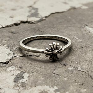 May include: A silver ring featuring a central cross-shaped emblem with rounded petals. The band is engraved with the text CHROME HEARTS and a small cross symbol on the inner surface. The metal has a polished finish with darkened recessed areas.