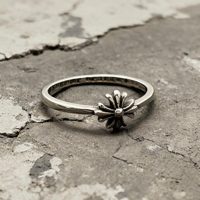 May include: A silver ring featuring a central cross-shaped emblem with rounded petals. The band is engraved with the text CHROME HEARTS and a small cross symbol on the inner surface. The metal has a polished finish with darkened recessed areas.
