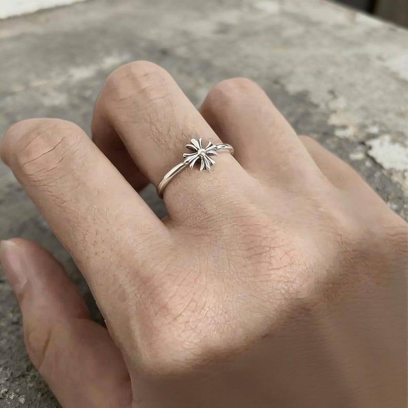 May include: A silver ring featuring a small, detailed cross emblem with four flared arms. The band is thin and polished, showcasing a metallic finish. The cross design is centred on the top of the ring, providing a clear view of the intricate metalwork.