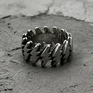 Pode incluir: Um anel de metal prateado com um design de banda cont&iacute;nua composto por formas de rel&acirc;mpagos repetidas e interconectadas. O anel tem um acabamento polido e reflexivo com &aacute;reas escuras rebaixadas entre cada rel&acirc;mpago, criando um alto contraste.