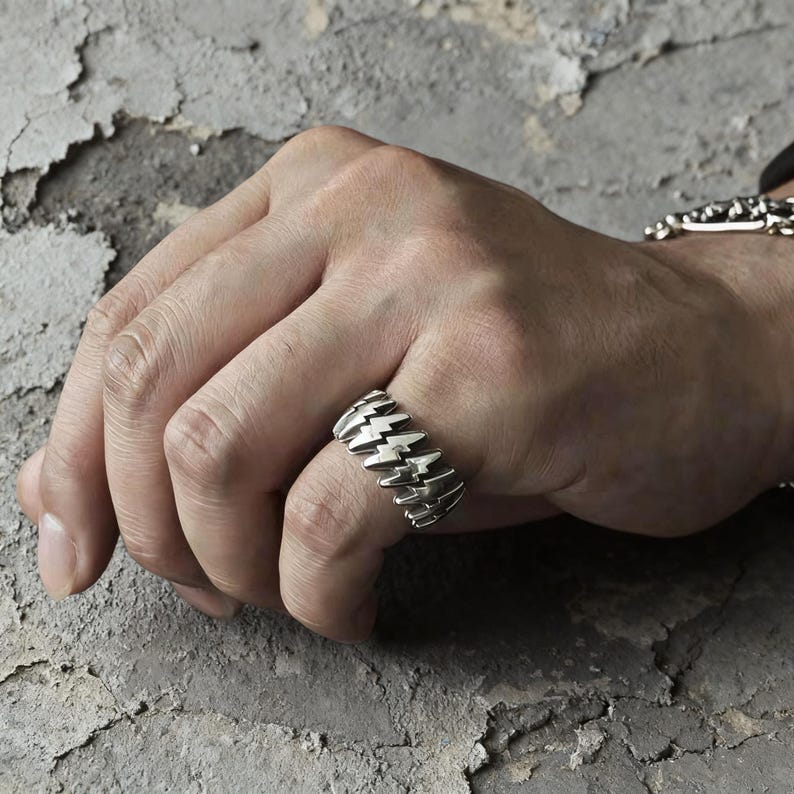 Pode incluir: Um anel de tom prateado com um padr&atilde;o repetitivo de formas de rel&acirc;mpagos em relevo ao redor de toda a banda. O metal tem um acabamento polido com &aacute;reas rebaixadas escurecidas que destacam o design afiado e angular dos rel&acirc;mpagos.