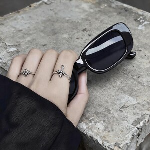 May include: Two silver rings featuring a detailed cross design worn on the fingers, paired with black rectangular sunglasses. The rings have a polished metallic finish with dark recessed detailing, while the sunglasses have a glossy black frame.