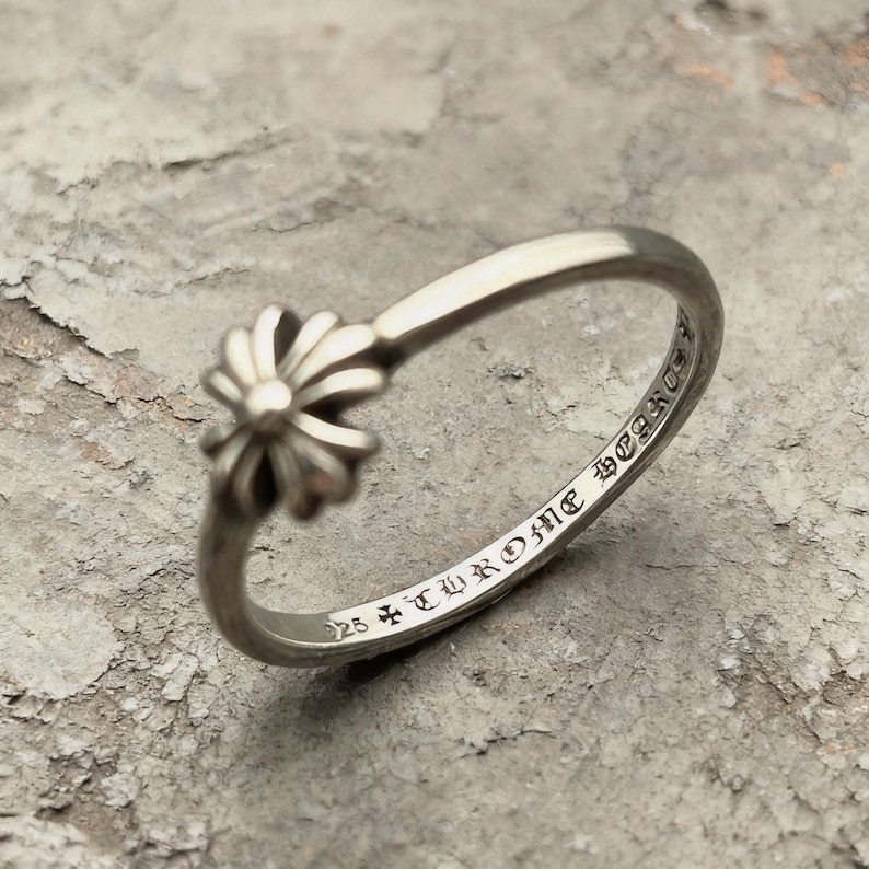 May include: A silver ring featuring a small, multi-petaled floral motif on the band. The interior of the band is engraved with the text 925 and Chrome Hearts in a gothic-style font, indicating the sterling silver composition of the jewellery piece.