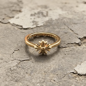 May include: A gold-toned ring featuring a central cross-shaped emblem with rounded, petal-like arms. The band is smooth and circular, with visible engraved text along the inner surface of the ring, showcasing a polished metallic finish throughout.