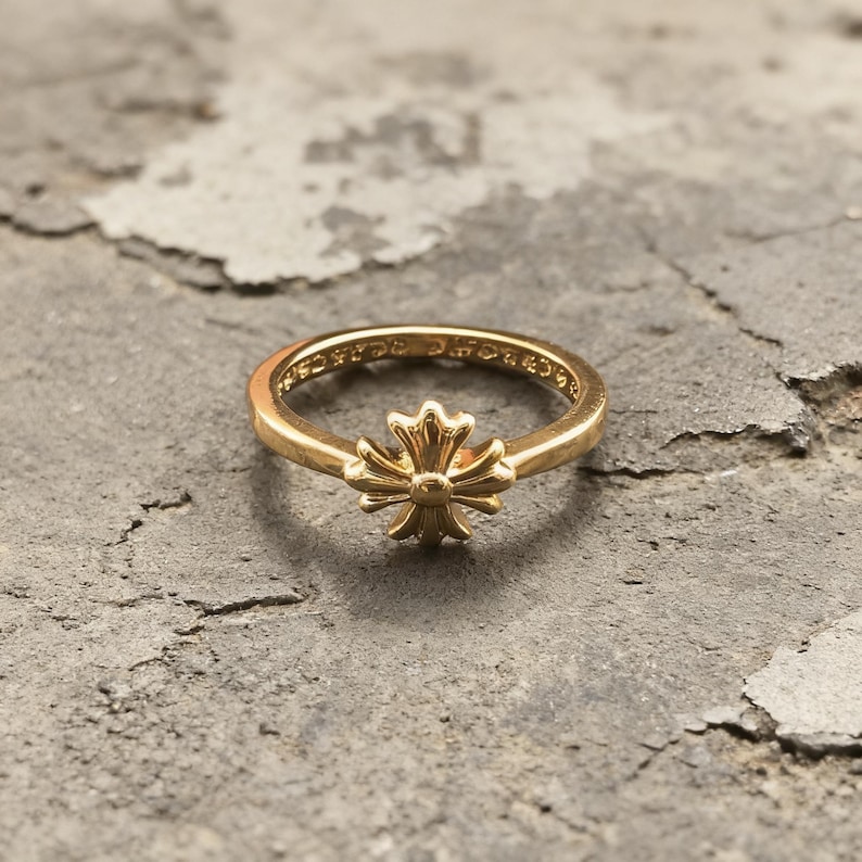 May include: A gold-toned ring featuring a central cross-shaped emblem with rounded, petal-like arms. The band is smooth and circular, with visible engraved text along the inner surface of the ring, showcasing a polished metallic finish throughout.