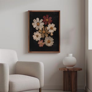 May include: A framed floral art print featuring a dark background with pressed flowers in shades of white, pink, and red. The flowers are arranged in a cluster, and the frame is made of wood. The artwork is displayed in a living room setting.