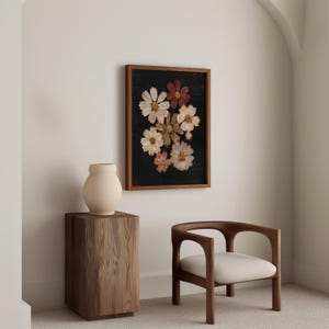 May include: A framed floral print featuring pressed flowers in shades of white, pink, and burgundy against a black background. The artwork is displayed in a room with a wooden side table, a cream-colored vase, and a wooden chair with a white cushion.