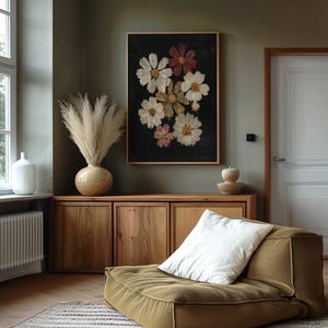 May include: A framed floral print featuring white, pink, and burgundy cosmos flowers against a dark background. The artwork is displayed above a wooden cabinet, with a vase of pampas grass and a modern olive green sofa in the room.