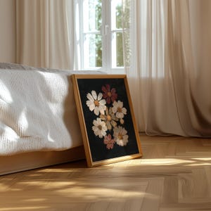 May include: A framed floral print featuring pressed cosmos flowers in shades of white, pink, and burgundy against a black background. The wooden frame is light-colored, and the artwork is leaning against a bed frame on a hardwood floor.