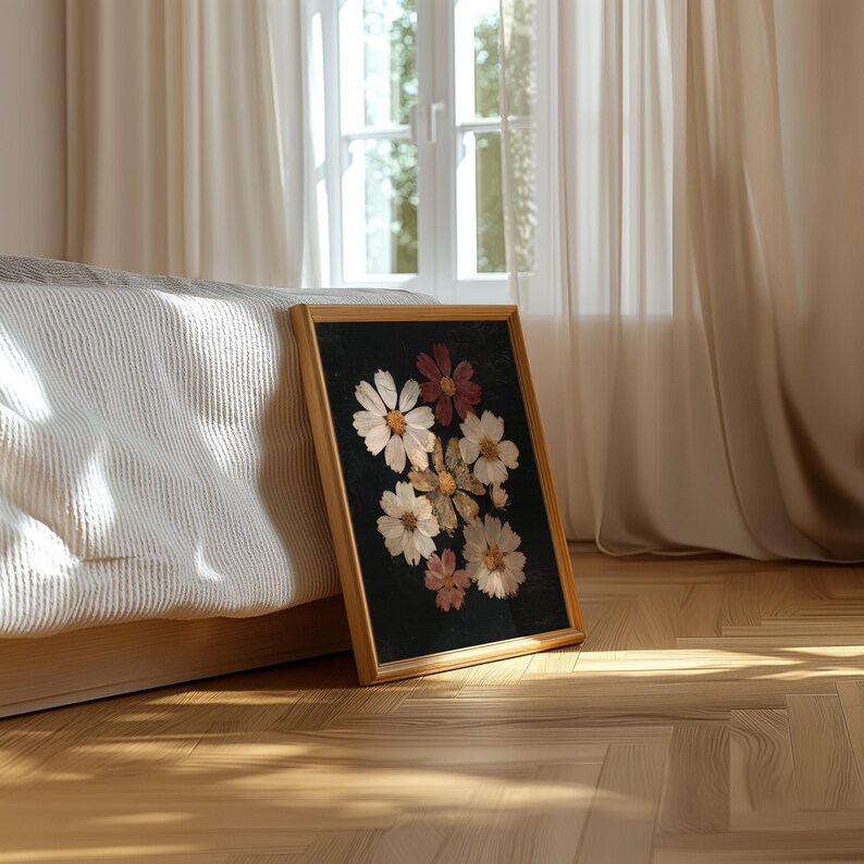 May include: A framed floral print featuring pressed cosmos flowers in shades of white, pink, and burgundy against a black background. The wooden frame is light-colored, and the artwork is leaning against a bed frame on a hardwood floor.