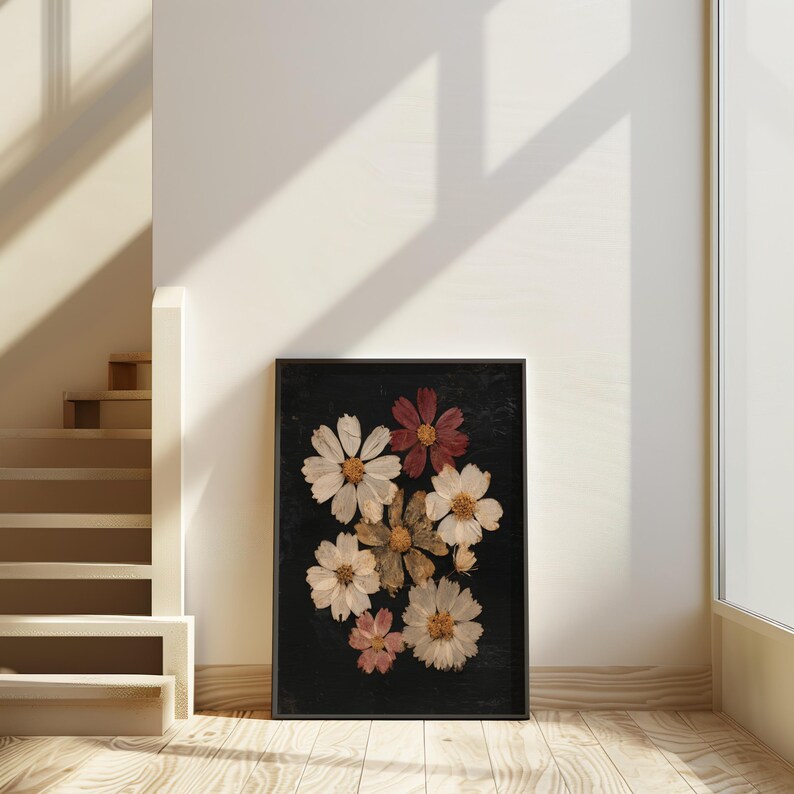 May include: A framed floral art piece featuring pressed flowers in shades of white, pink, and burgundy against a black background. The arrangement includes several daisy-like blooms. The artwork is displayed on a wooden floor.