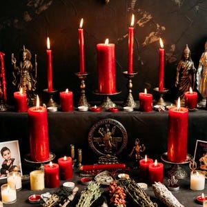 May include: A dark, moody scene featuring lit red candles of varying sizes, displayed on ornate candle holders. The arrangement includes small statues, dried herbs, and a decorative medallion, creating a mystical ambiance. The background is a dark, textured wall.