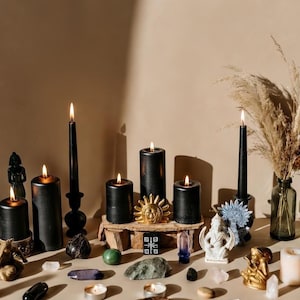 May include: An assortment of black candles, crystals, and decorative objects arranged on a beige surface. The candles vary in size and shape, some are lit. Other items include a sun-shaped ornament, small statues, and stones, creating a mystical theme.