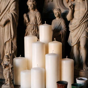 Pode incluir: Uma cole&ccedil;&atilde;o de velas de pilar de cor marfim de v&aacute;rias alturas, dispostas sobre uma superf&iacute;cie de madeira escura. As velas t&ecirc;m pavios vis&iacute;veis. Est&aacute;tuas antigas de figuras cl&aacute;ssicas est&atilde;o no fundo, juntamente com pequenos vasos decorativos.
