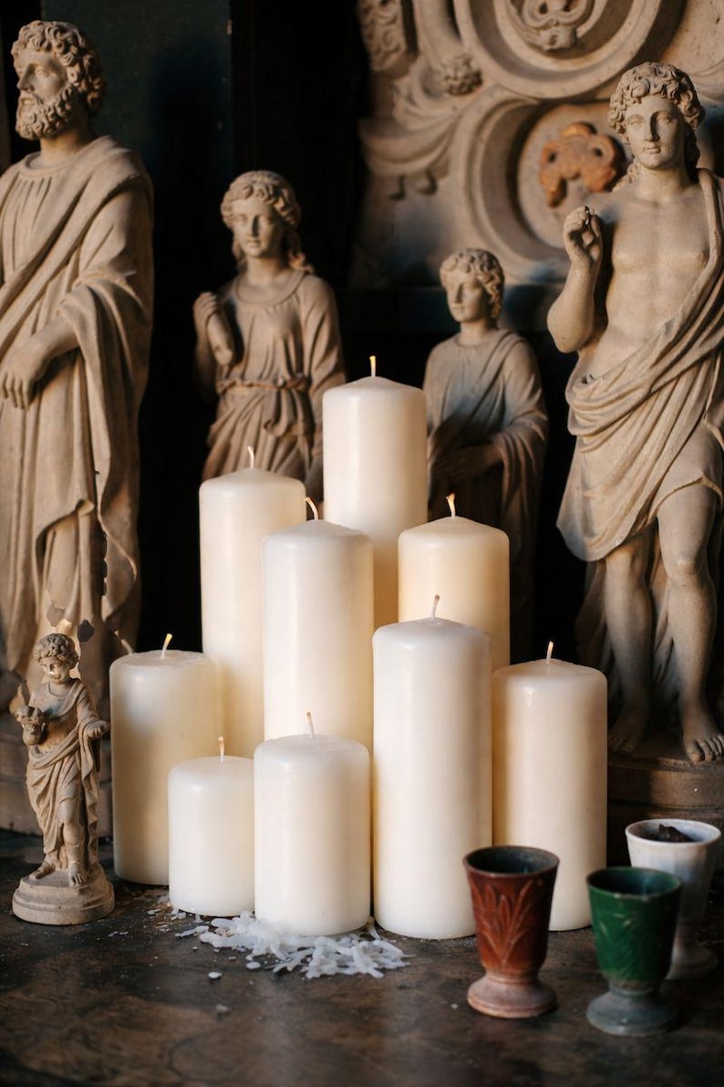 Pode incluir: Uma cole&ccedil;&atilde;o de velas de pilar de cor marfim de v&aacute;rias alturas, dispostas sobre uma superf&iacute;cie de madeira escura. As velas t&ecirc;m pavios vis&iacute;veis. Est&aacute;tuas antigas de figuras cl&aacute;ssicas est&atilde;o no fundo, juntamente com pequenos vasos decorativos.
