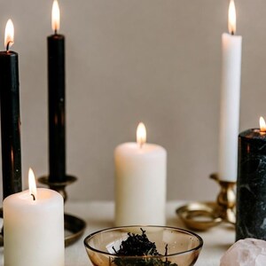 May include: An assortment of lit candles in varying sizes and colors, including black, white, and marble-patterned. A small glass bowl holds dried herbs, and a few crystals are scattered around. The scene evokes a sense of ambiance and spirituality.