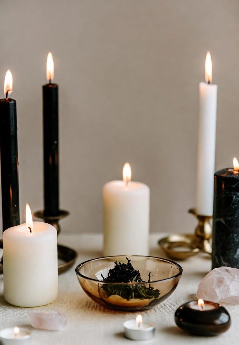 May include: An assortment of lit candles in varying sizes and colors, including black, white, and marble-patterned. A small glass bowl holds dried herbs, and a few crystals are scattered around. The scene evokes a sense of ambiance and spirituality.