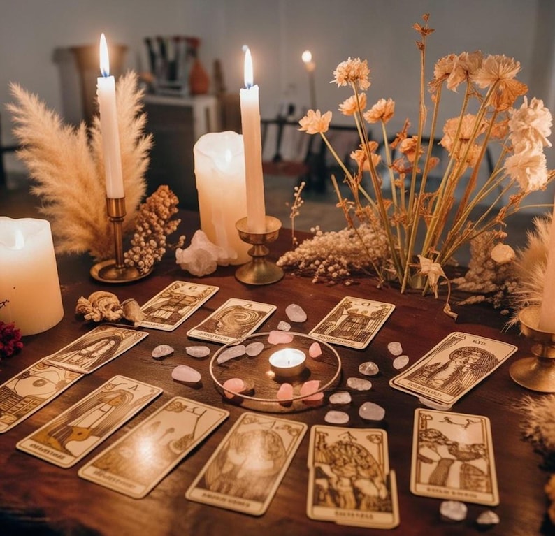 Pode incluir: Uma mesa de madeira exibe uma configura&ccedil;&atilde;o de leitura de cartas de tar&ocirc;. Velas acesas em casti&ccedil;ais de lat&atilde;o e uma vela central iluminam a cena. As cartas de tar&ocirc; s&atilde;o dispostas com cristais de quartzo rosa espalhados e flores secas, criando um ambiente m&iacute;stico.