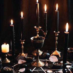 May include: A mystical scene featuring lit black candles in ornate holders, a chalice, and small bowls with herbs and crystals arranged on a black surface. A white pentagram is drawn on the surface, creating a dark, gothic aesthetic.