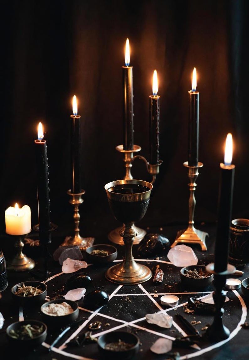 May include: A mystical scene featuring lit black candles in ornate holders, a chalice, and small bowls with herbs and crystals arranged on a black surface. A white pentagram is drawn on the surface, creating a dark, gothic aesthetic.