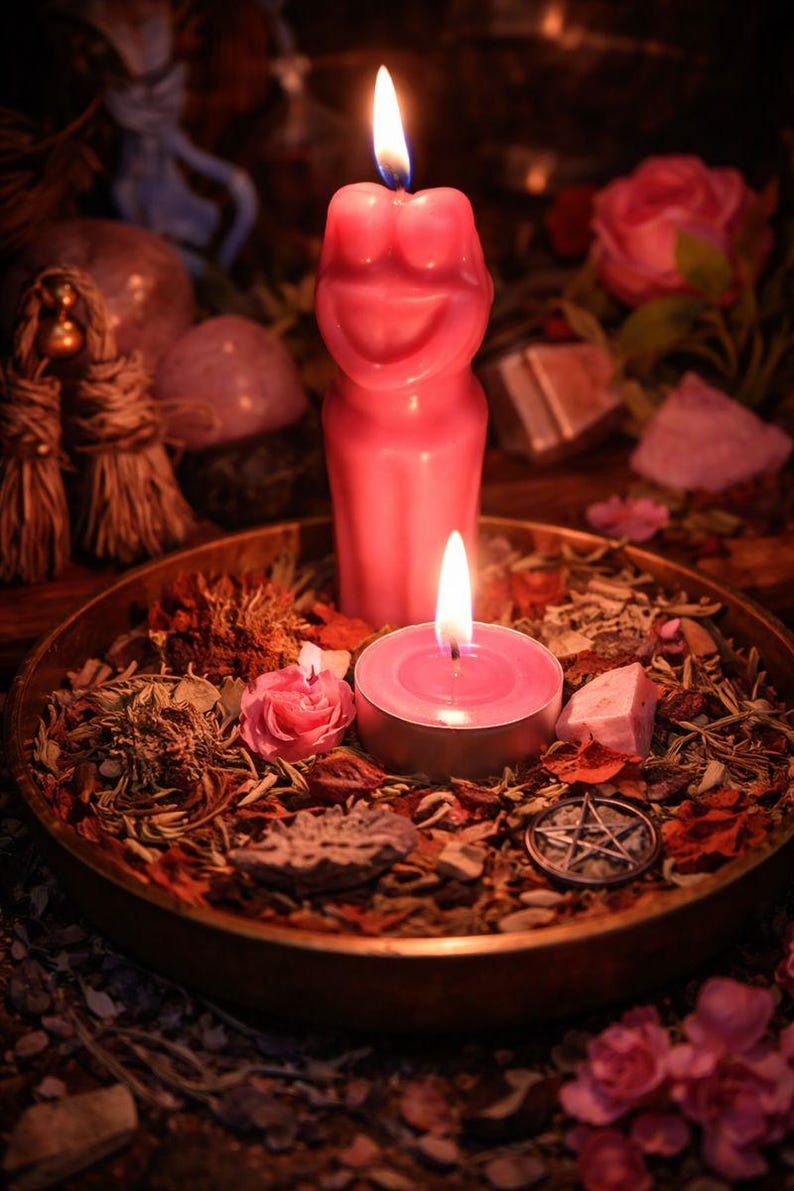 May include: A pink figure candle and a small round candle, both lit with flames. They sit in a brass bowl with herbs, flowers, and a pentagram charm. The scene is set with a mystical theme.