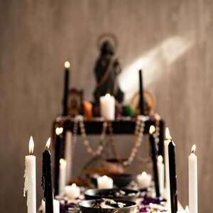 May include: A table setting with lit candles, bowls, and decorative elements. Tall black and white candles are interspersed with smaller tea lights. Bowls contain dark contents, and purple flower petals are scattered around. A statue and beads are on a shelf in the background.