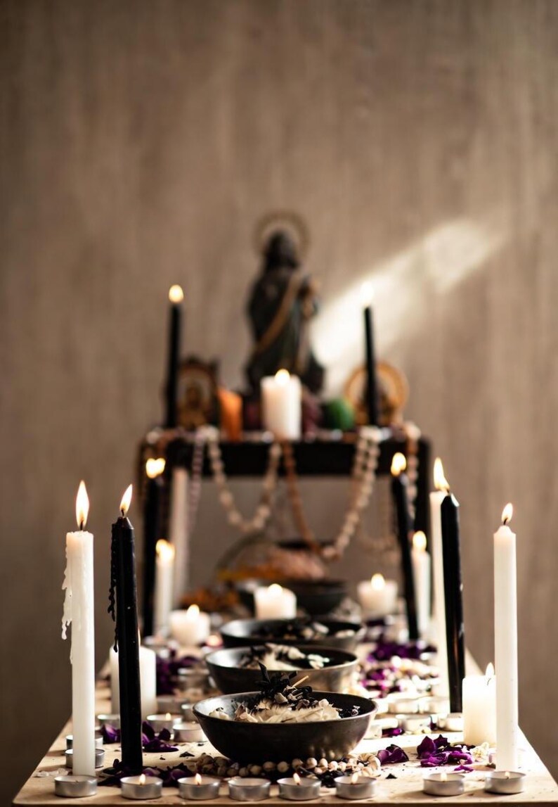 May include: A table setting with lit candles, bowls, and decorative elements. Tall black and white candles are interspersed with smaller tea lights. Bowls contain dark contents, and purple flower petals are scattered around. A statue and beads are on a shelf in the background.