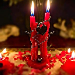 May include: Two red candles are joined by a decorative chain and a black heart-shaped accent. The candles are lit with blue and yellow flames, and surrounded by melted wax and rose petals. The overall theme is romantic or spiritual.
