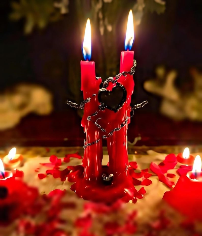 May include: Two red candles are joined by a decorative chain and a black heart-shaped accent. The candles are lit with blue and yellow flames, and surrounded by melted wax and rose petals. The overall theme is romantic or spiritual.
