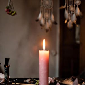 May include: A lit pink candle on a black surface, surrounded by crystals and feathers. Dream catchers hang in the background, creating a mystical atmosphere. The candle flame is bright. The scene evokes a sense of spirituality.