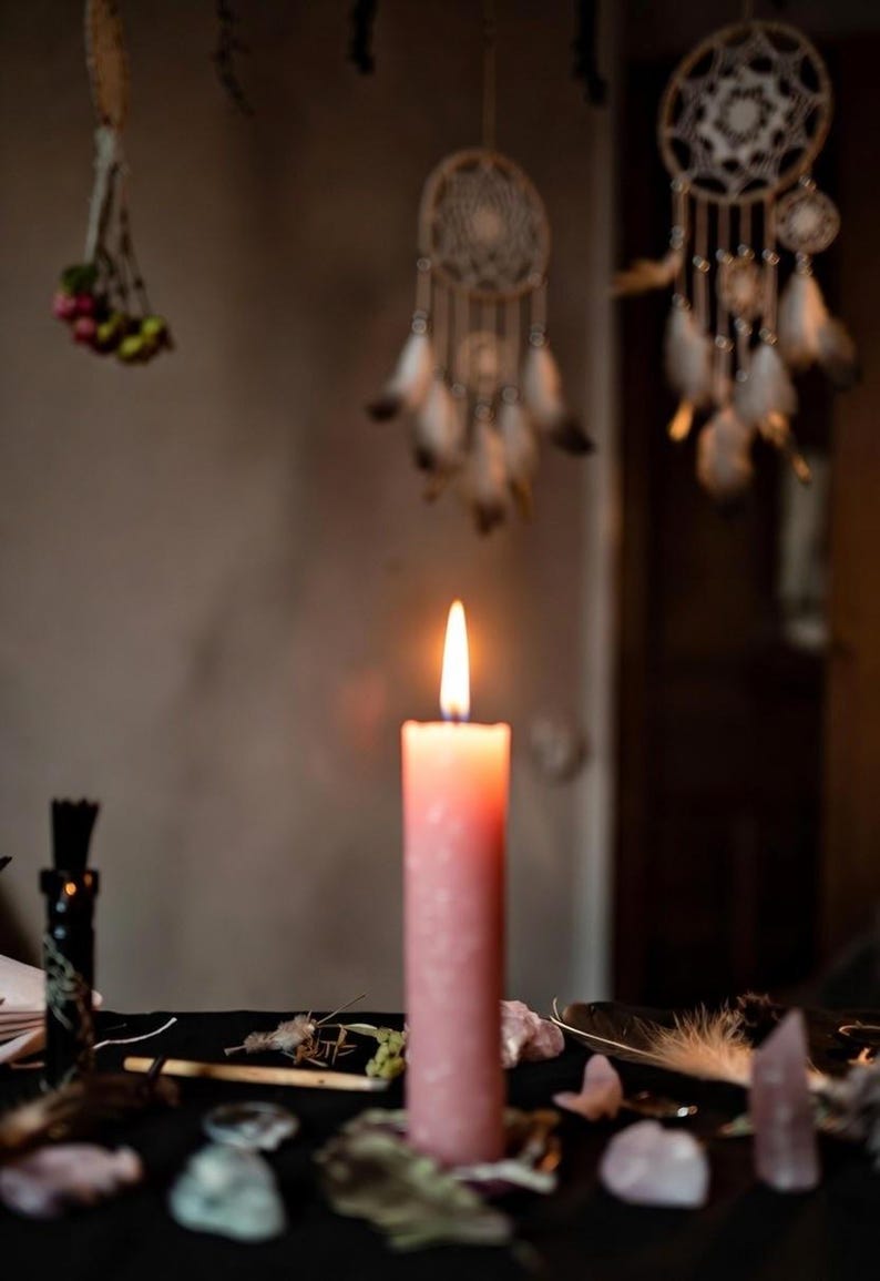 May include: A lit pink candle on a black surface, surrounded by crystals and feathers. Dream catchers hang in the background, creating a mystical atmosphere. The candle flame is bright. The scene evokes a sense of spirituality.